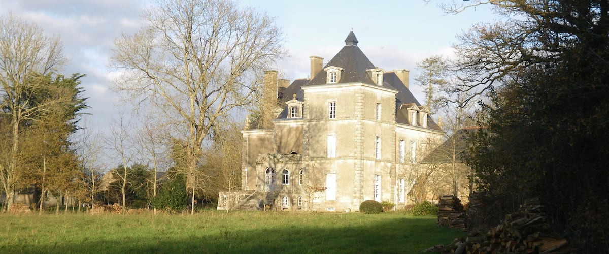 A large, historic stone building with a prominent tower and multiple windows, surrounded by a grassy field and trees.