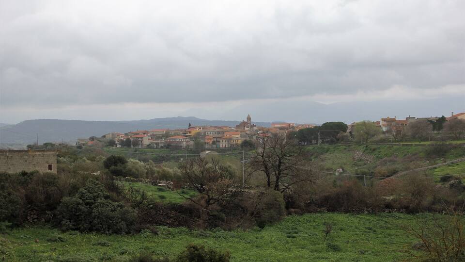 A town with red-roofed buildings on a hillside, surrounded by greenery and hills in the background.