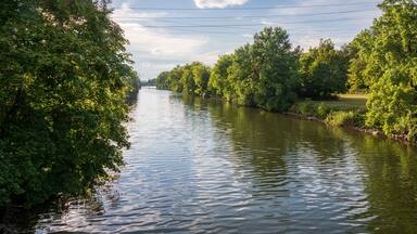 A calm river flanked by lush green trees under a partly cloudy sky.