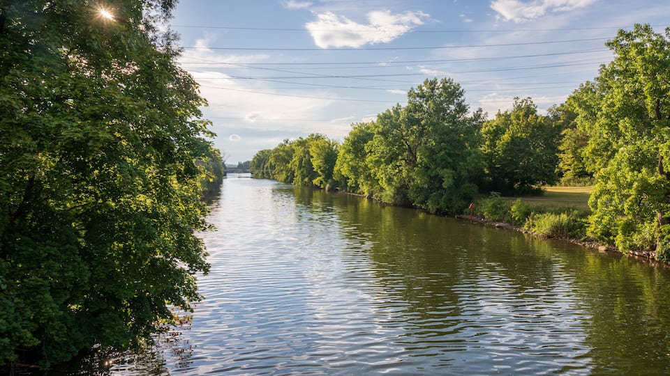 A calm river flanked by lush green trees under a partly cloudy sky.