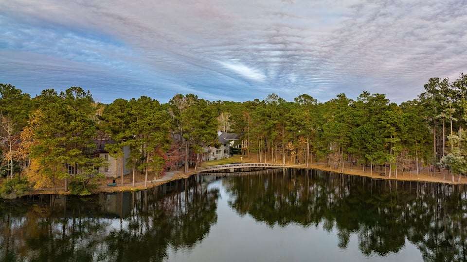 A calm lake with a wooden bridge, surrounded by dense trees and a house in the background.