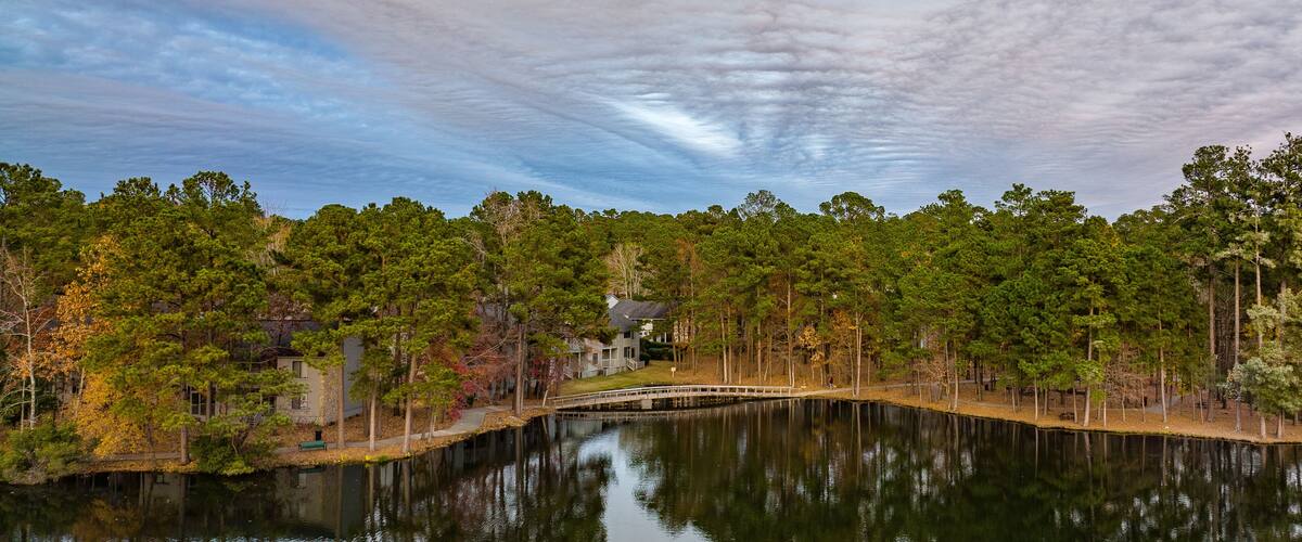 A calm lake with a wooden bridge, surrounded by dense trees and a house in the background.