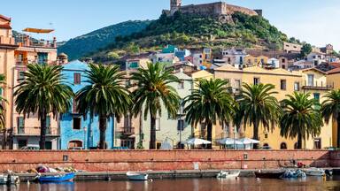 A coastal town with colorful buildings, palm trees, and a historic castle on a hill.