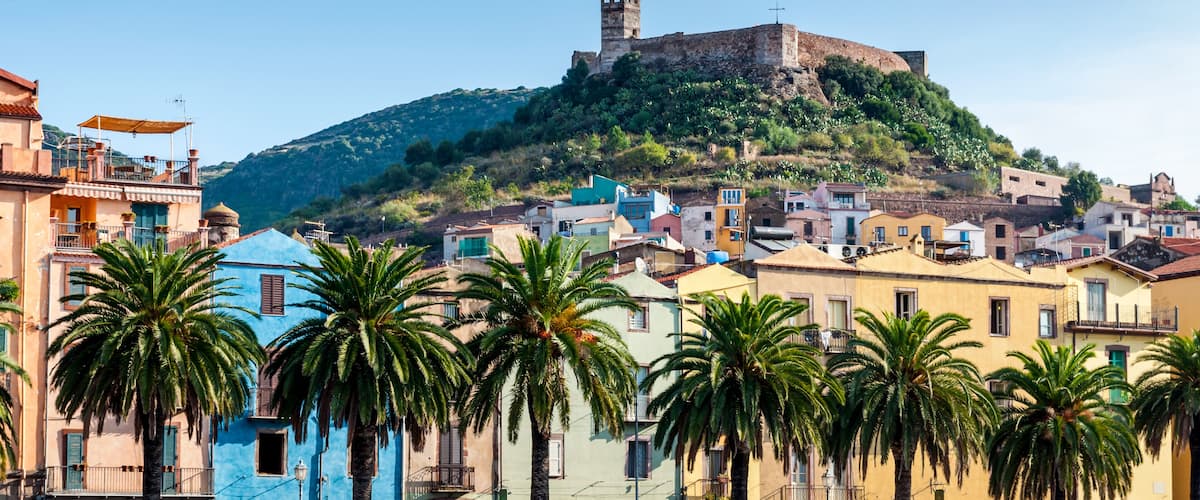 A coastal town with colorful buildings, palm trees, and a historic castle on a hill.