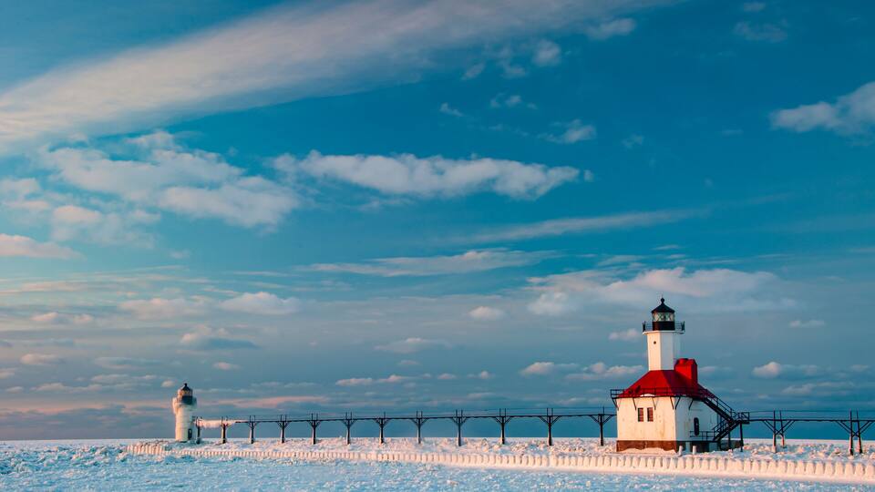 A lighthouse with a red roof and white walls, surrounded by snow-covered ground and a clear blue sky with clouds.