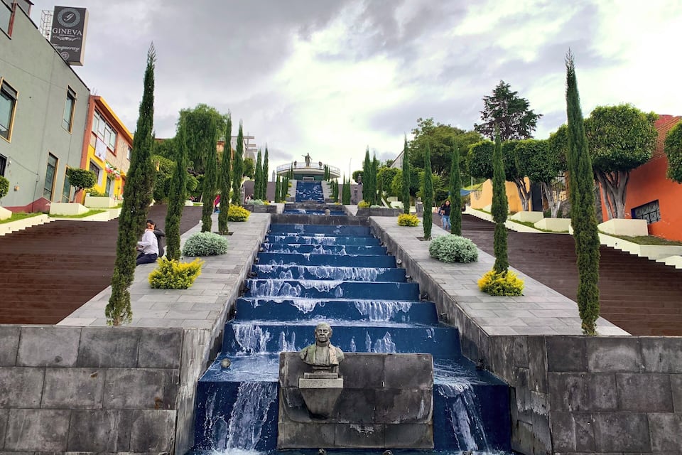 A cascading water feature with a statue in the center, flanked by neatly trimmed trees and buildings.