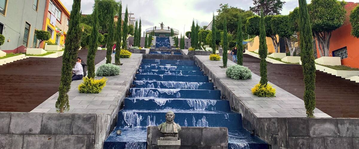 A cascading water feature with a statue in the center, flanked by neatly trimmed trees and buildings.