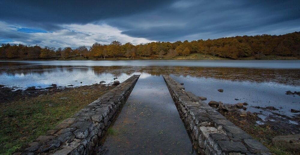 A stone pathway leading to a body of water with autumn foliage in the background.