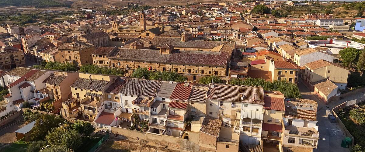A historic town with terracotta-roofed buildings, a prominent church, and rolling hills in the background.