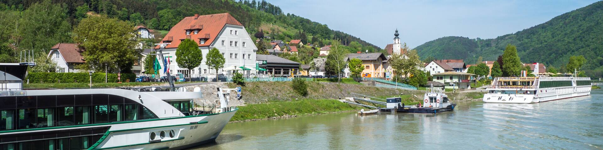 A river with boats, a town with houses and a church, and green hills in the background.