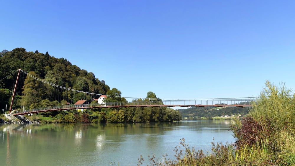 A suspension bridge over a calm river, surrounded by lush greenery and hills.