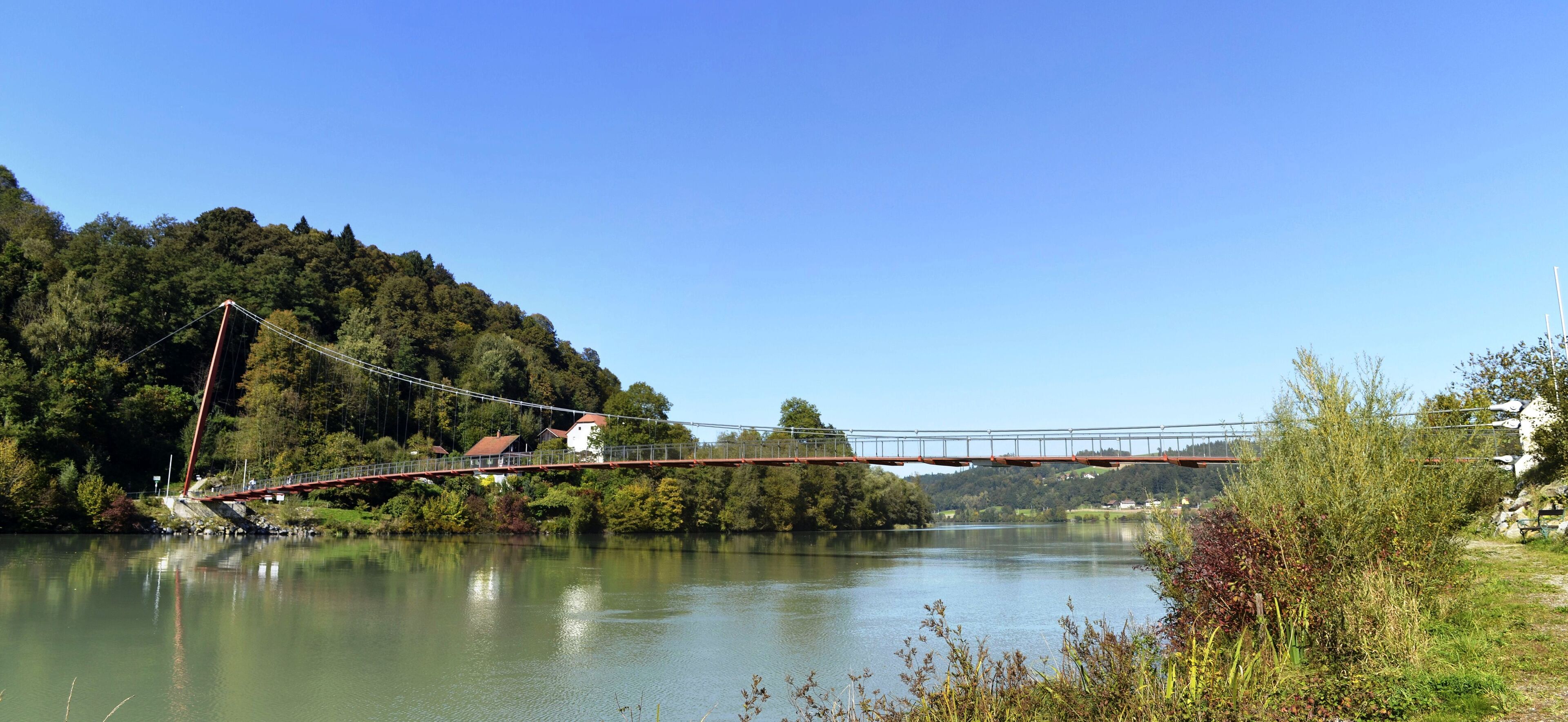 A suspension bridge over a calm river, surrounded by lush greenery and hills.