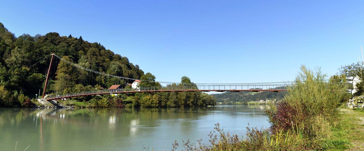 A suspension bridge over a calm river, surrounded by lush greenery and hills.