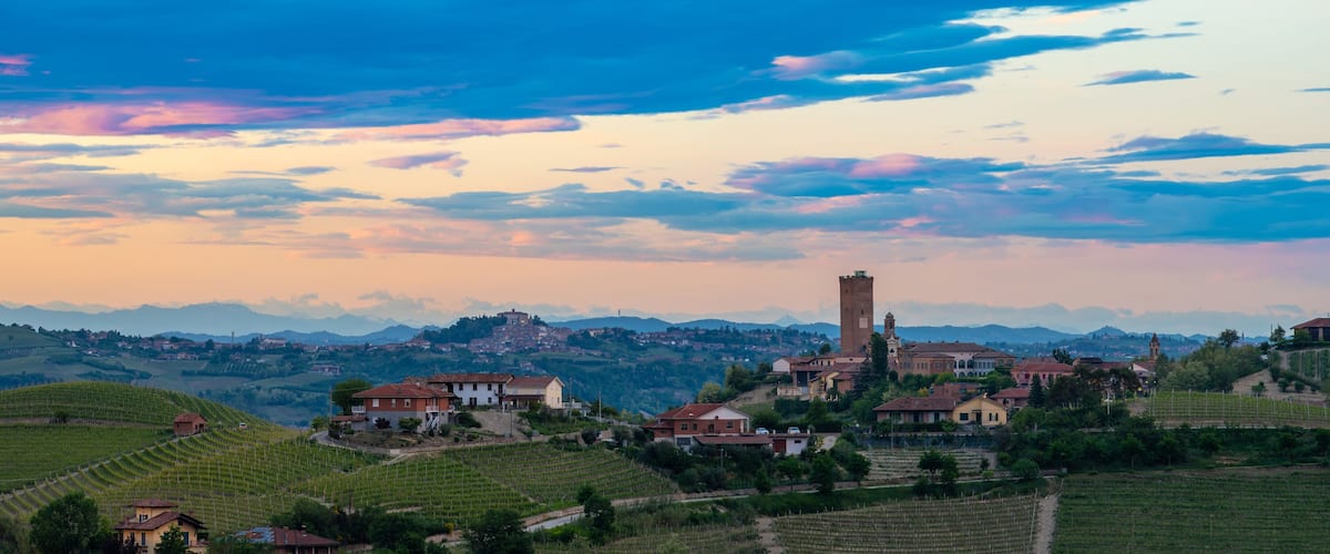 A hillside village with vineyards, a prominent tower, and a mountain range in the background.