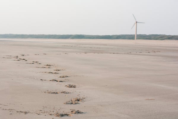 A sandy beach with footprints leading towards a wind turbine in the distance.