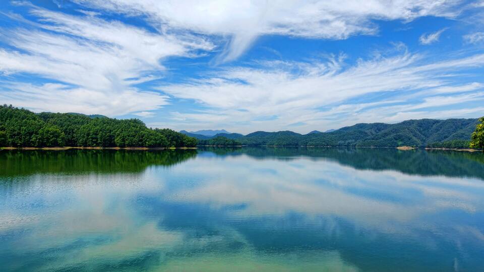 A calm lake with clear reflections, surrounded by dense green forest and distant mountains under a partly cloudy sky.