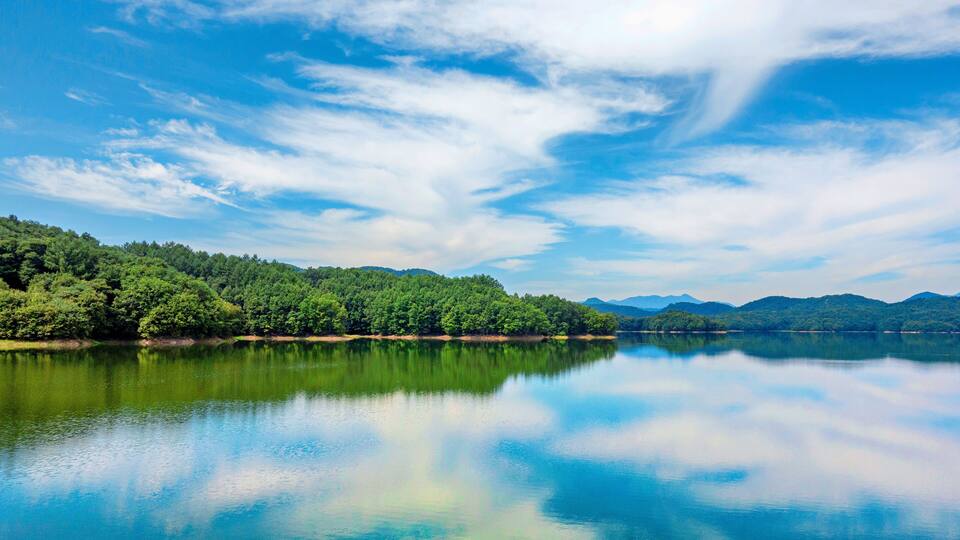 A calm lake with a forested shoreline and distant mountains under a partly cloudy sky.