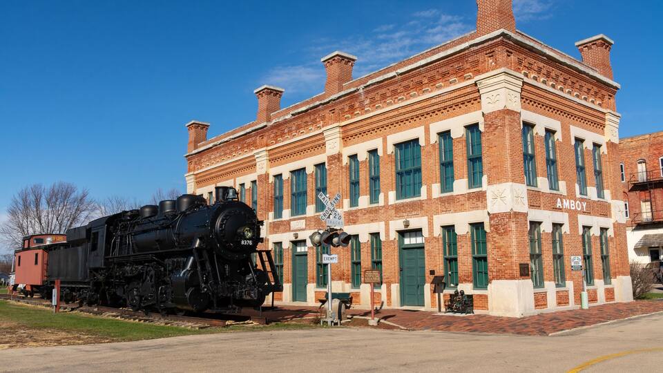 A historic train station with a black steam locomotive and a brick building labeled "AMBOY".