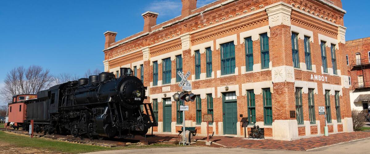 A historic train station with a black steam locomotive and a brick building labeled "AMBOY".