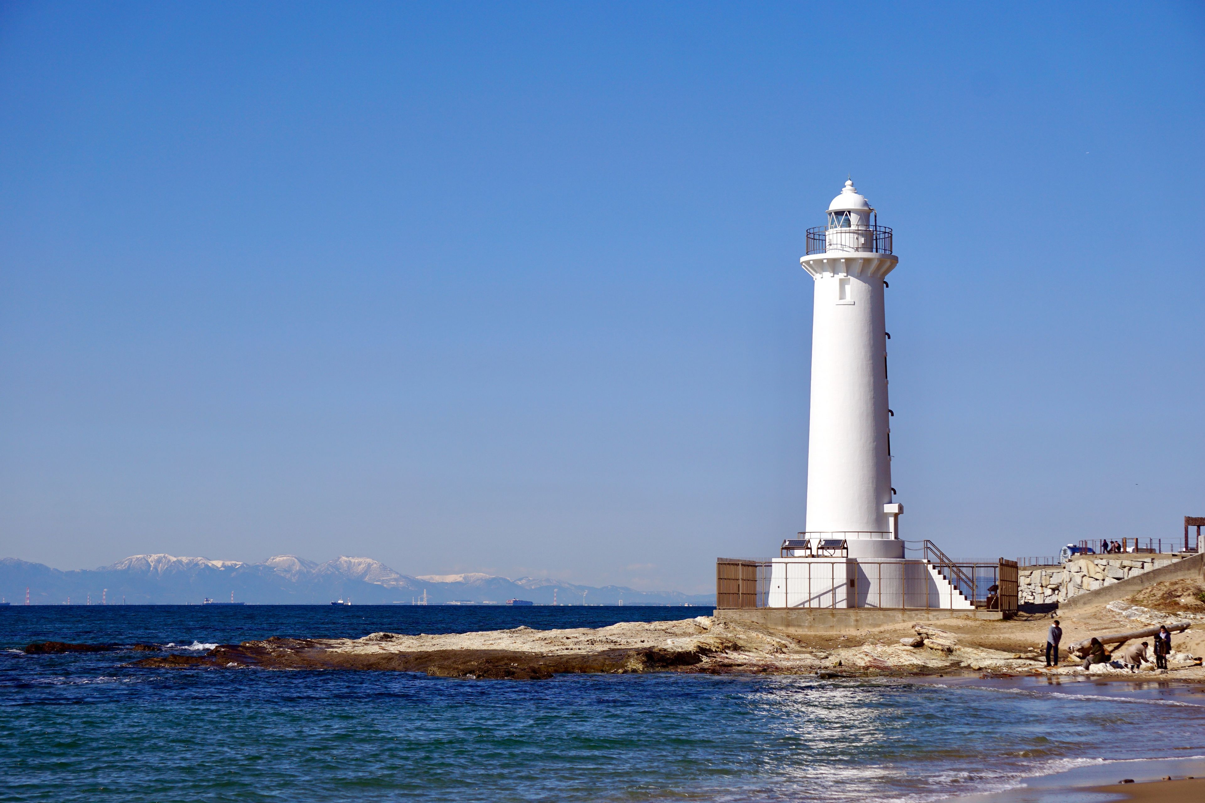 The historic white Noma Lighthouse against a blue sky and distant snowy mountains in Aichi, Japan.