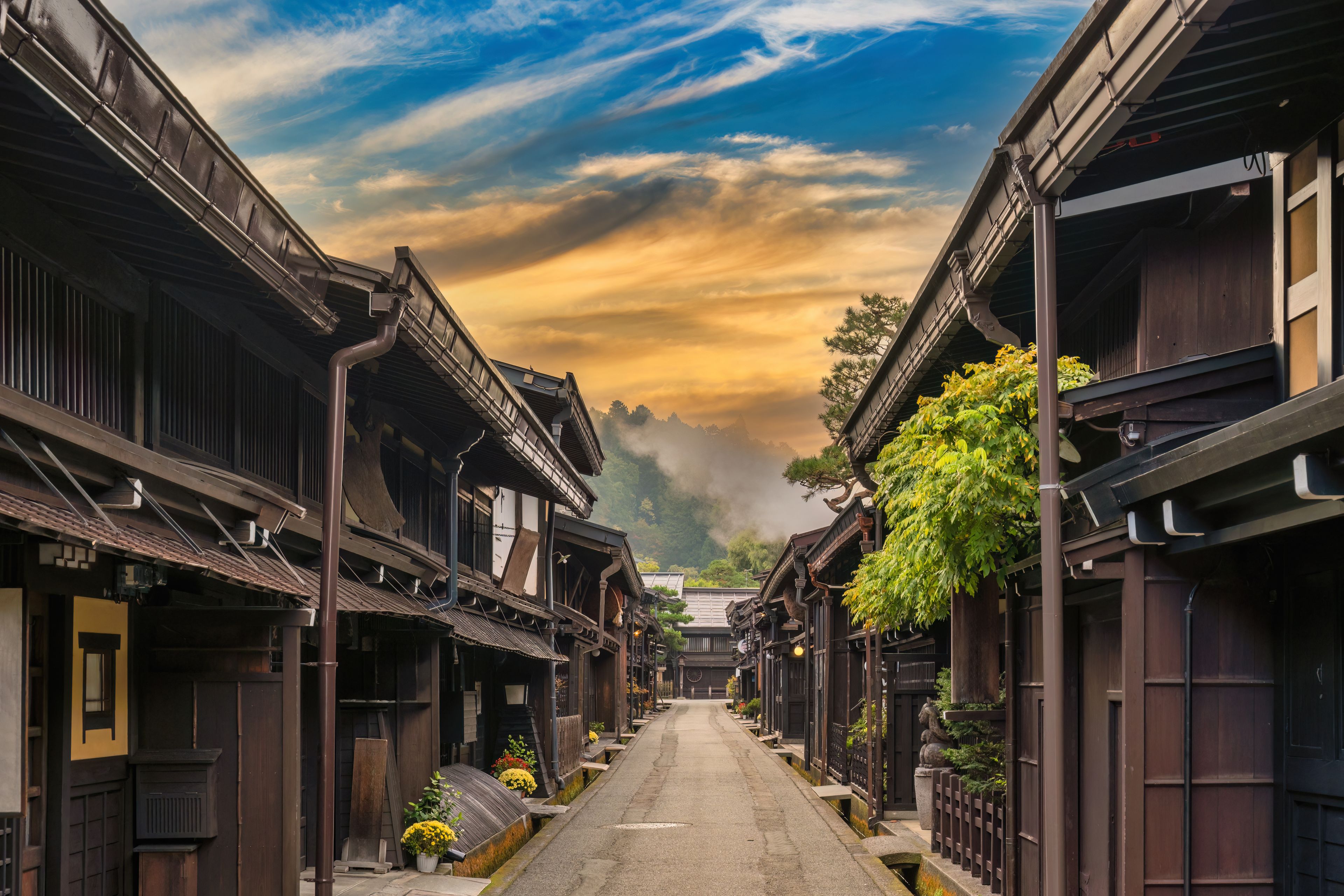 A narrow street lined with traditional wooden buildings, a clear sky with clouds, and a distant mountain range.