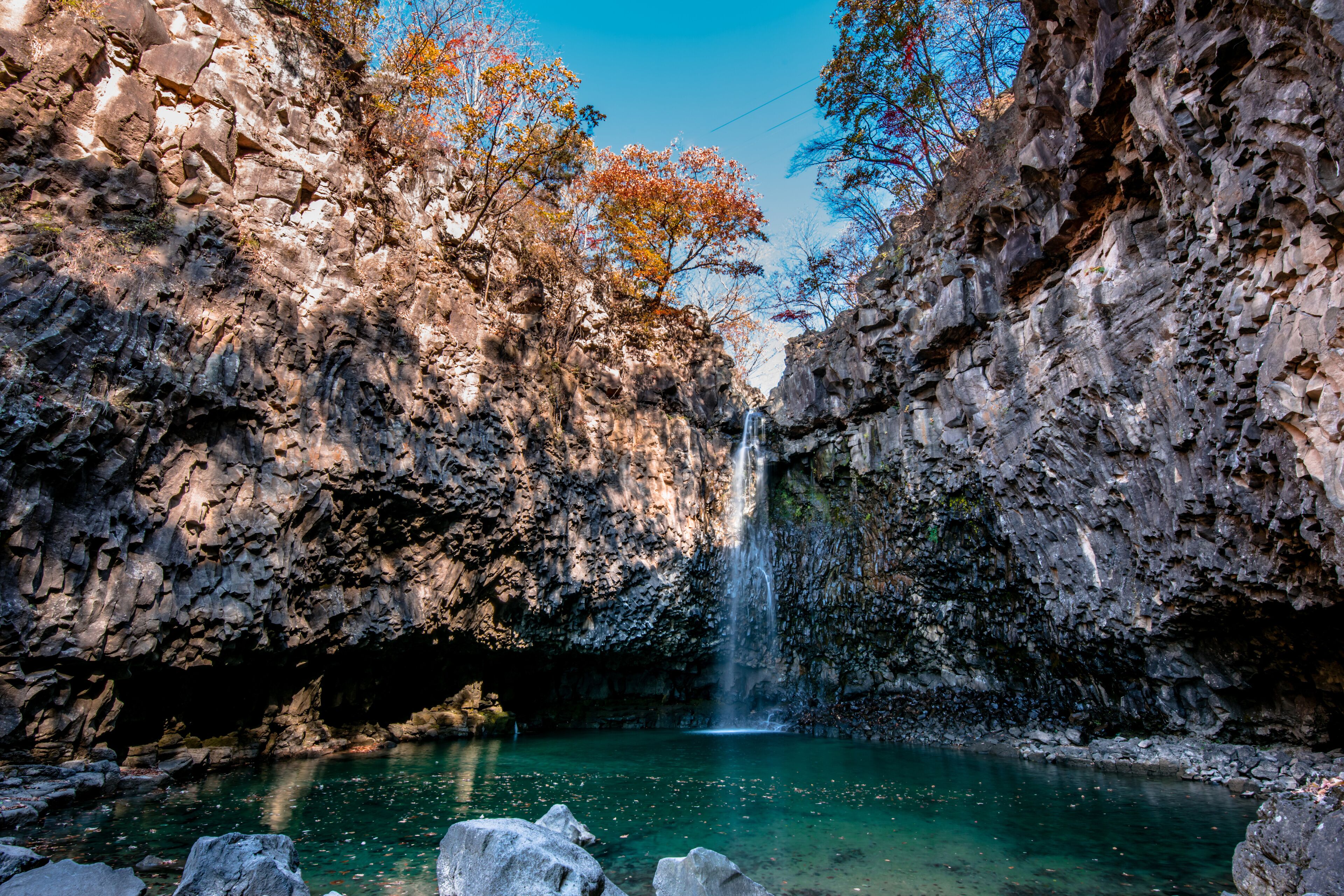 A waterfall cascading into a clear pool surrounded by rocky cliffs and autumn-colored trees.