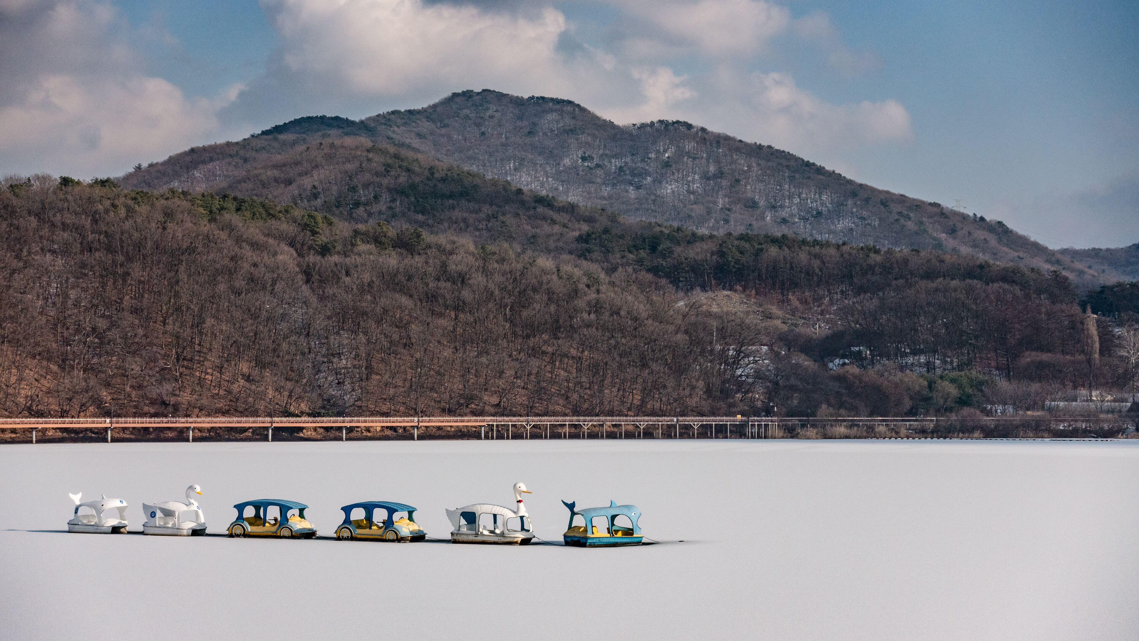 A frozen lake with a row of colorful boats and a bridge in the background.