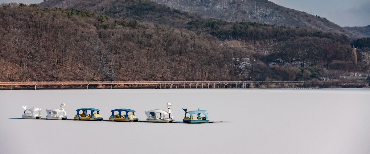 A frozen lake with a row of colorful boats and a bridge in the background.