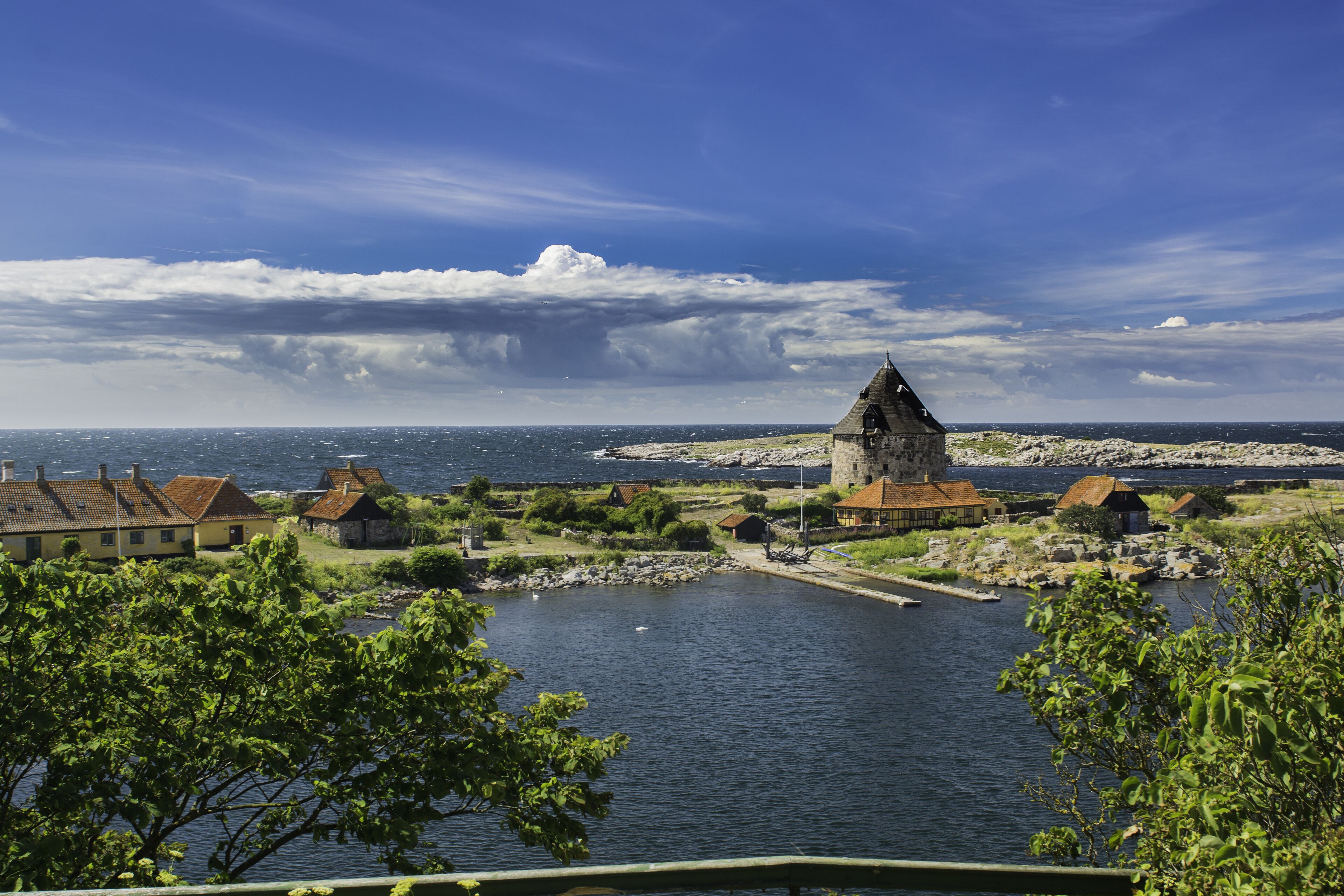 A coastal village with a prominent tower, houses, and a calm sea under a partly cloudy sky.