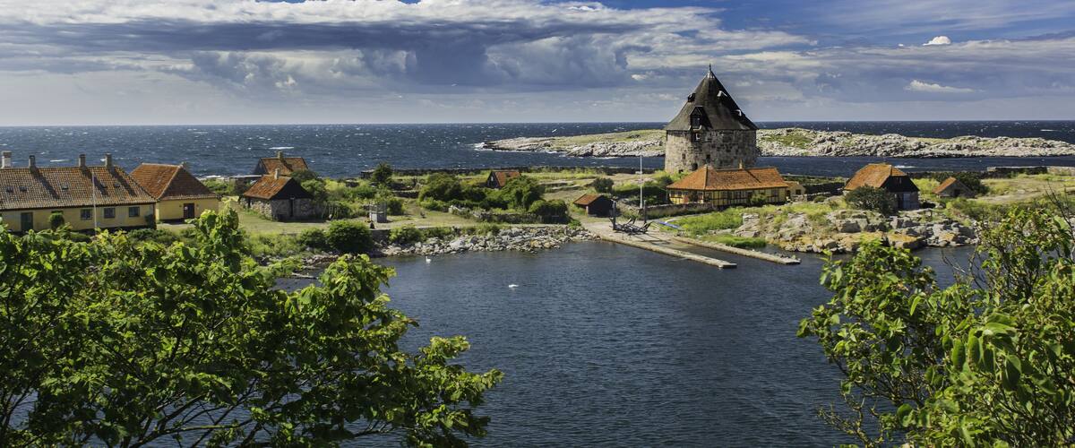 A coastal village with a prominent tower, houses, and a calm sea under a partly cloudy sky.