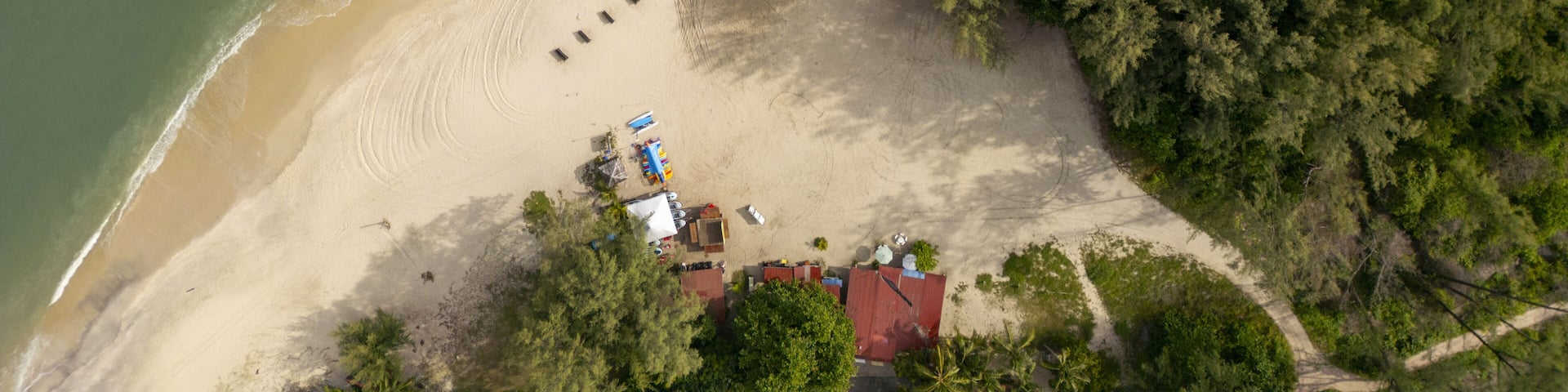 An aerial view of a beach with a row of beach huts, a small structure, and dense forested area.