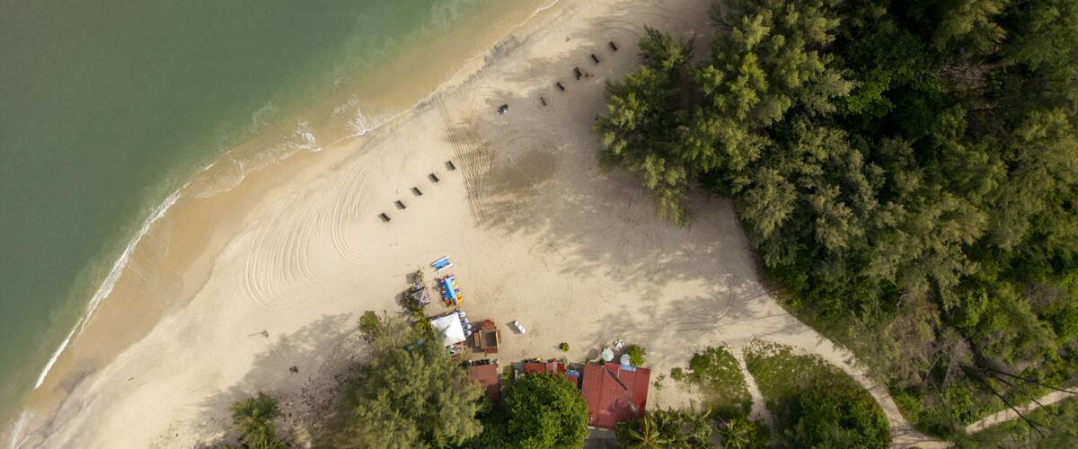 An aerial view of a beach with a row of beach huts, a small structure, and dense forested area.