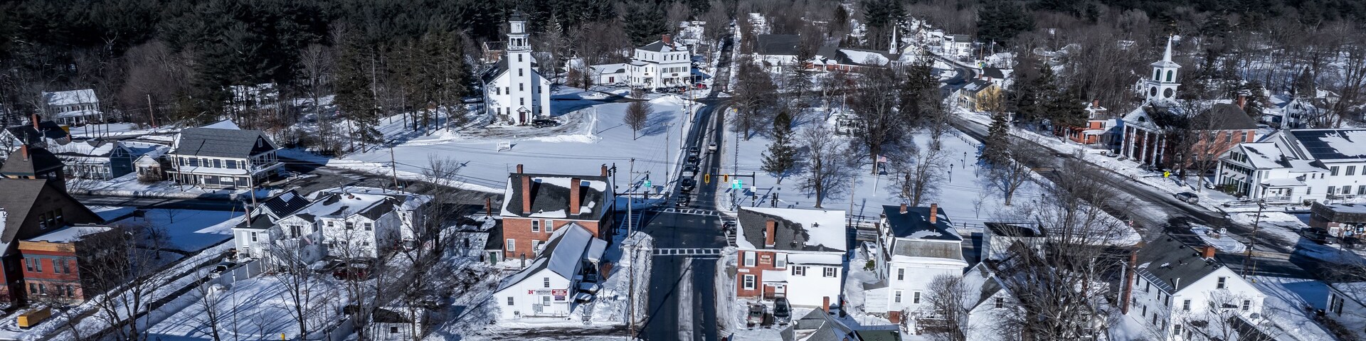 Un pueblo cubierto de nieve con un campanario de iglesia destacado, un puente y una combinación de edificios residenciales y comerciales.