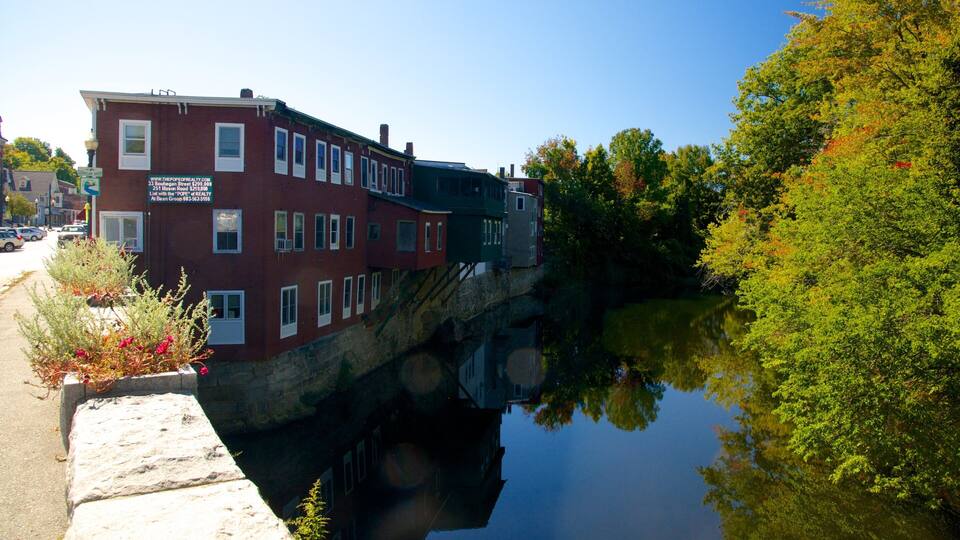A red brick building with a sign, a stone ledge, and a calm river reflecting trees.