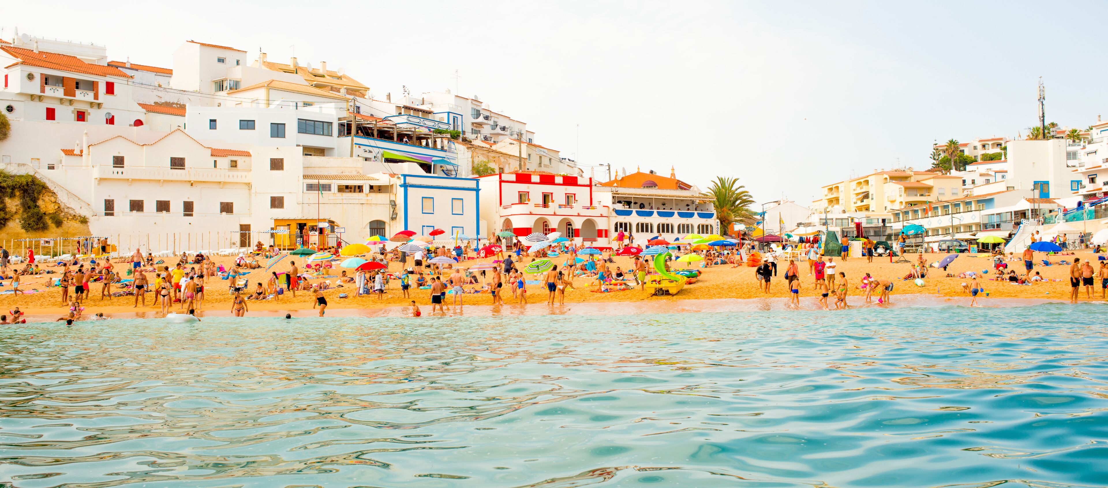 A crowded beach with people, umbrellas, and buildings in the background.