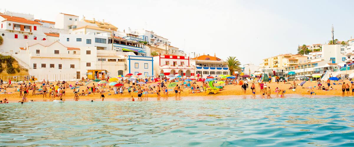 A crowded beach with people, umbrellas, and buildings in the background.