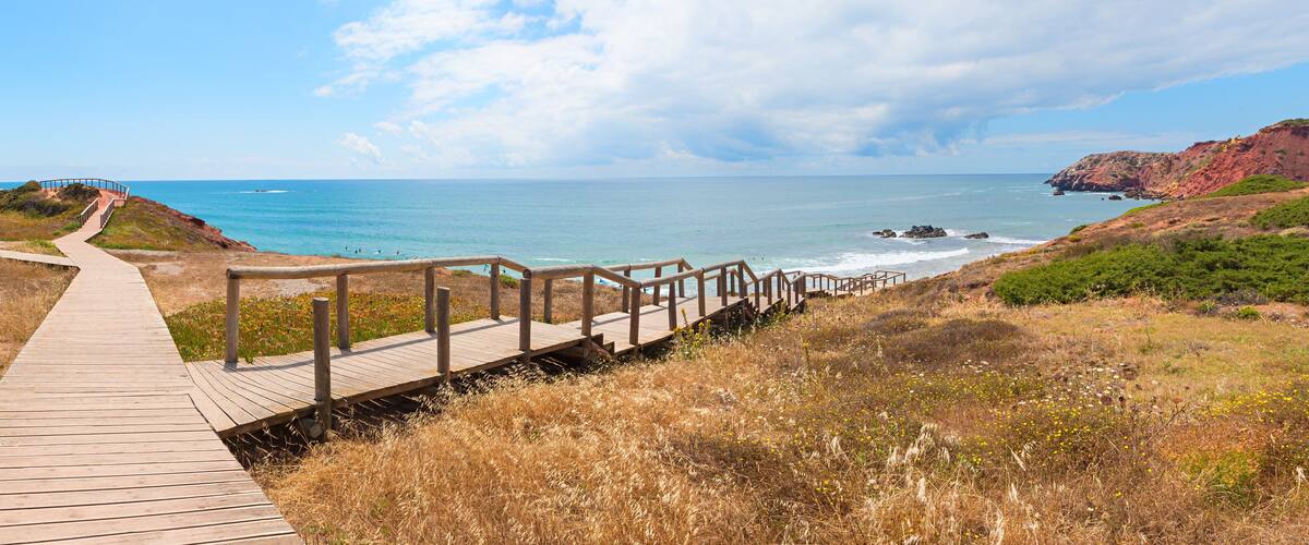 A wooden boardwalk leading to a beach with a clear view of the ocean and distant hills.