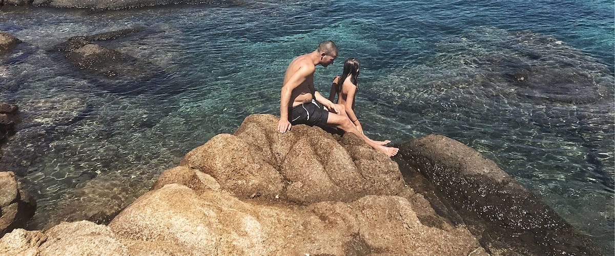 A couple sitting on rocks by a clear blue sea with a hilly landscape in the background.