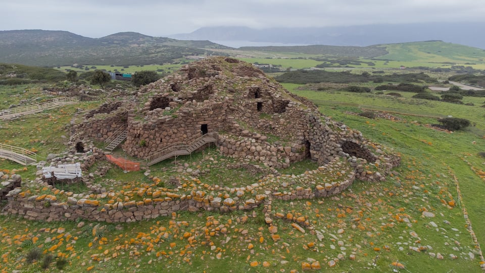 A stone ruin with a circular wall and a staircase leading up to it, surrounded by green fields and mountains in the background.
