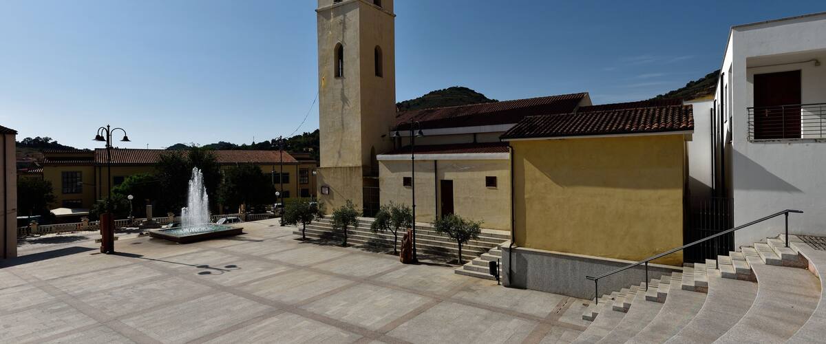 A square with a fountain, a tall tower, and a building with a red-tiled roof.