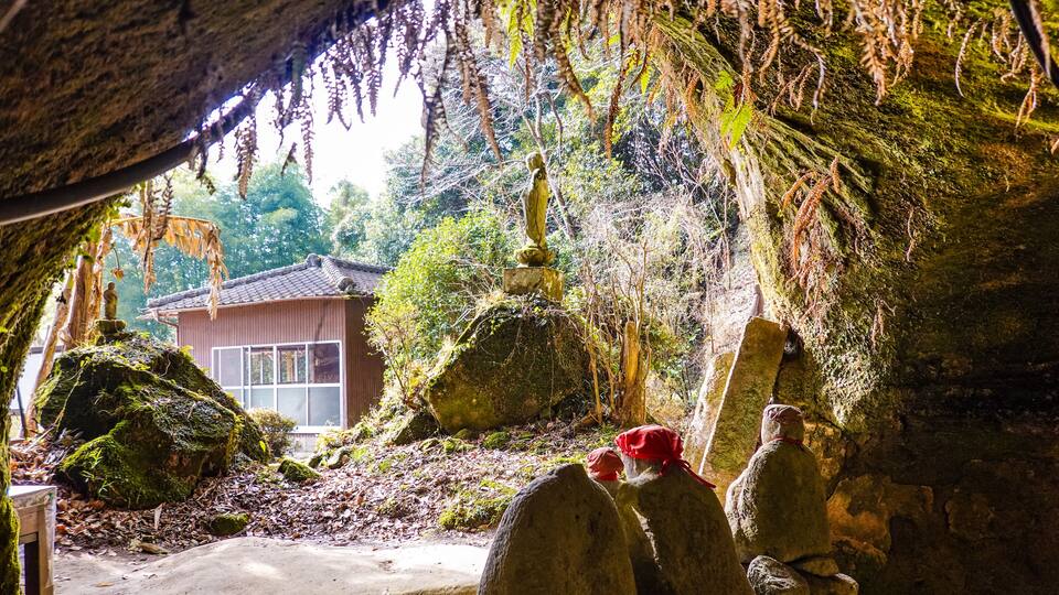 A cave entrance with moss-covered rocks and a traditional building in the background.
