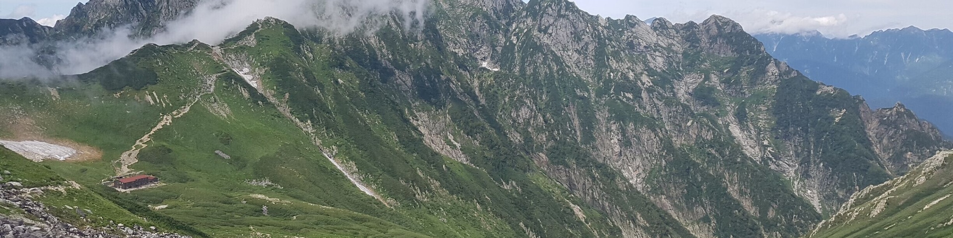 Uma cordilheira com um pico proeminente parcialmente coberto por nuvens, vales verdes exuberantes e um pequeno edifício com telhado vermelho.