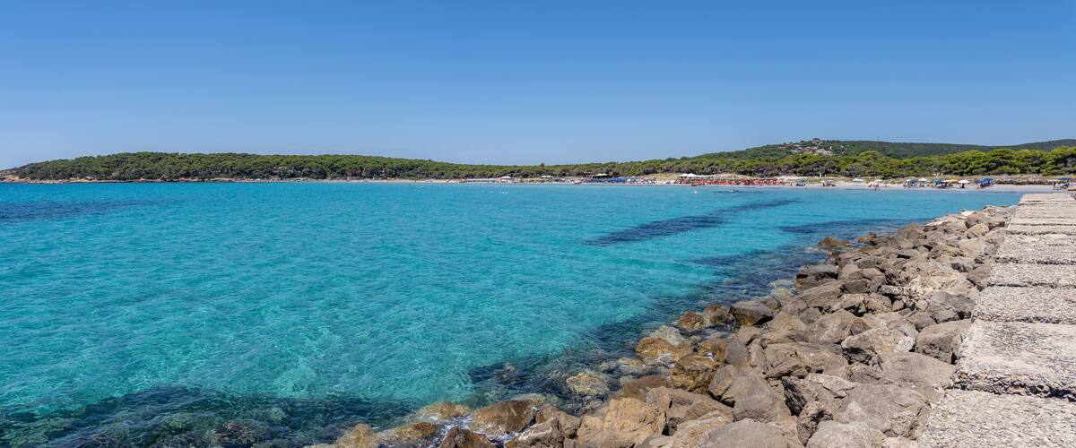 A coastal scene with clear turquoise waters, a rocky shoreline, and a distant hill with trees.