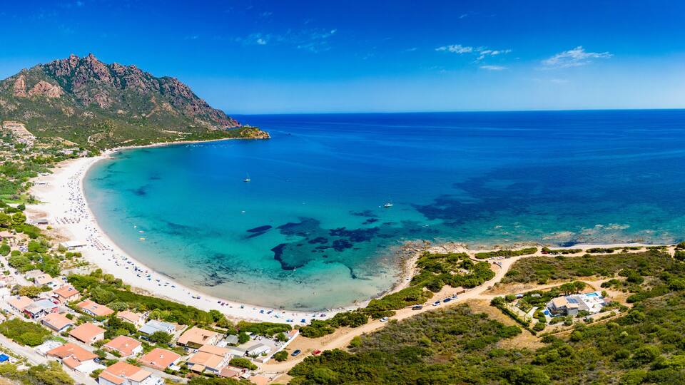A coastal town with a beach, clear blue waters, and a mountain in the background.