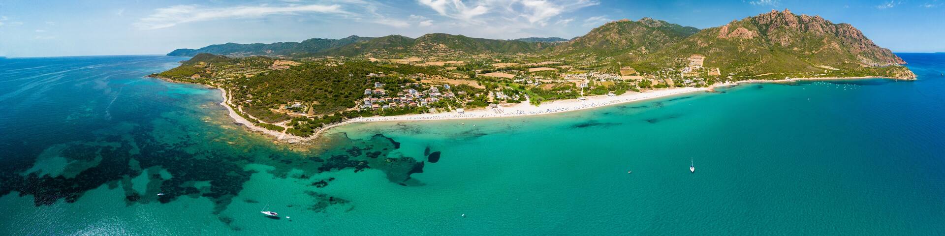 An aerial view of a coastal island with a beach, clear turquoise waters, and a mountainous landscape.
