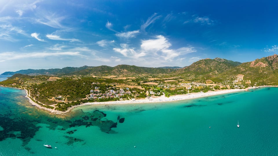 An aerial view of a coastal island with a beach, clear turquoise waters, and a mountainous landscape.