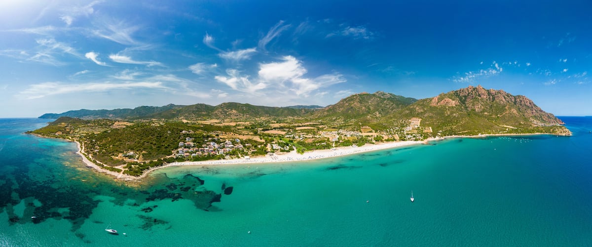 An aerial view of a coastal island with a beach, clear turquoise waters, and a mountainous landscape.