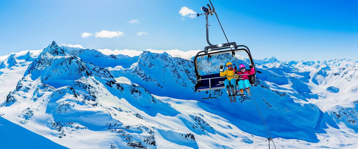 A ski lift with passengers in colorful winter gear, surrounded by snow-covered mountains and a clear blue sky.