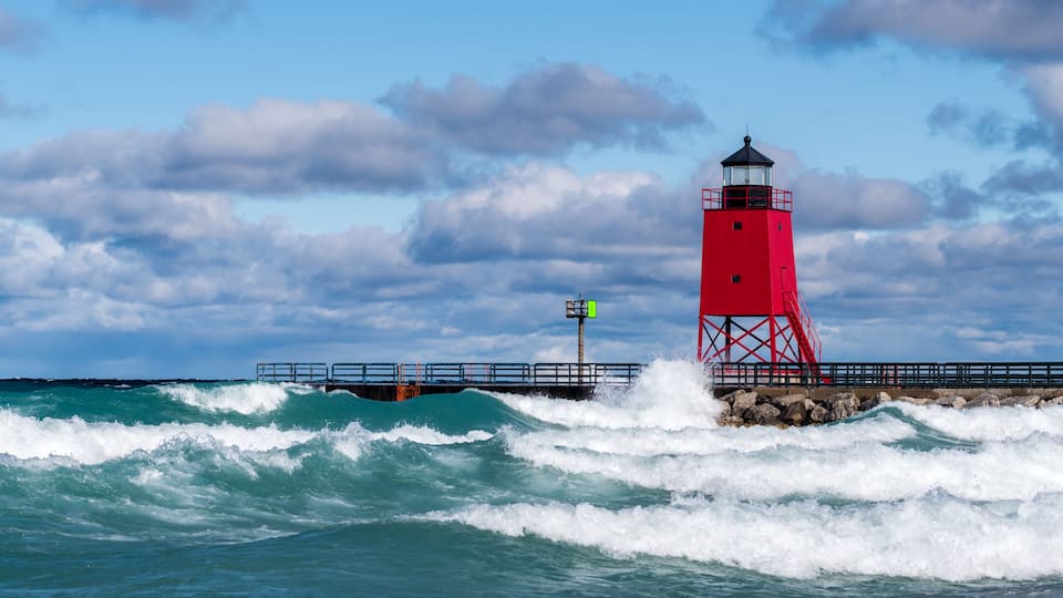 A red lighthouse on a rocky outcrop with waves crashing against it.
