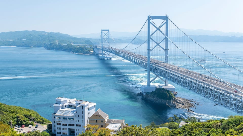 A large suspension bridge spanning a body of water, with a mountainous landscape in the background.